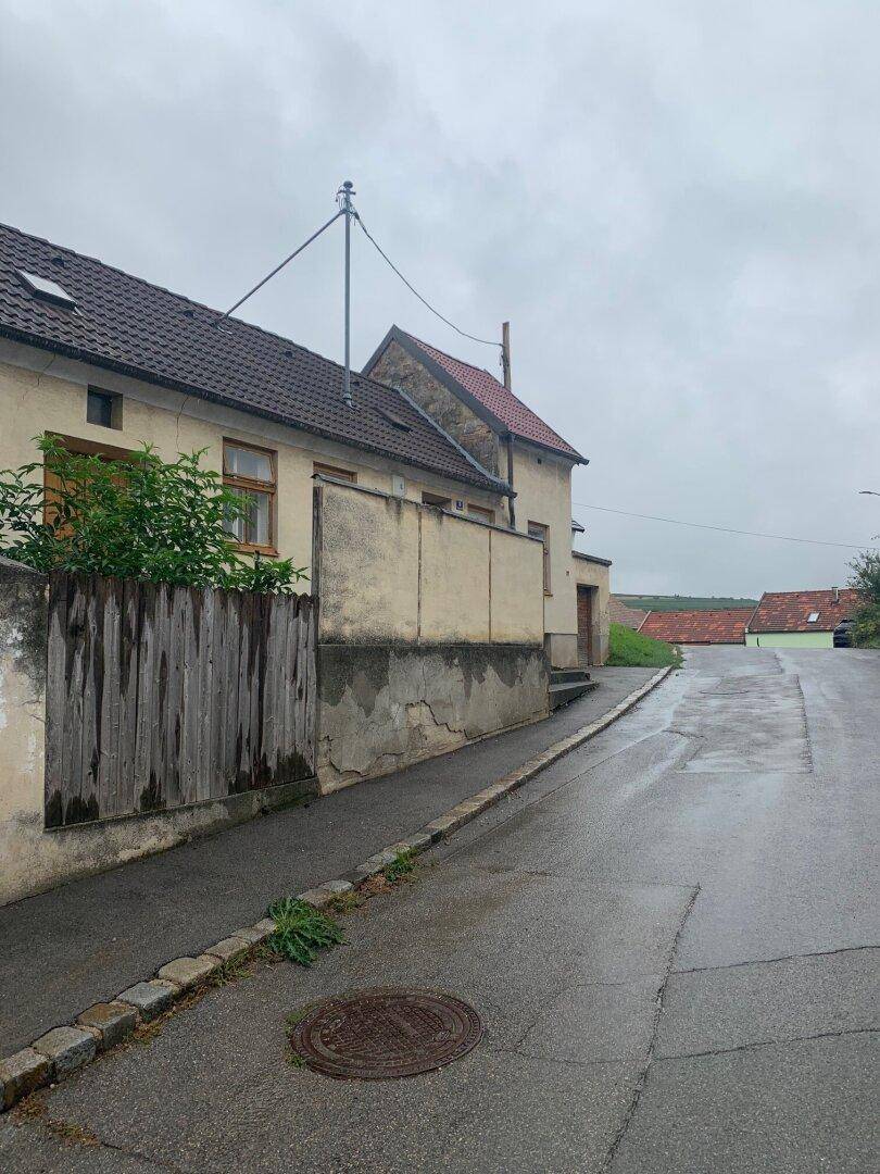 Seitenansicht eines älteren Hauses mit Putzfassade und Holzzaun an einer leicht ansteigenden, nassen Straße.