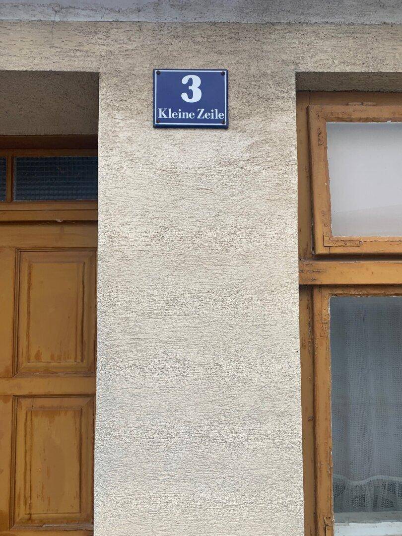 Nahaufnahme der Hausnummer 3 'Kleine Zeile' an der Putzfassade neben der Holztür und einem Fenster.