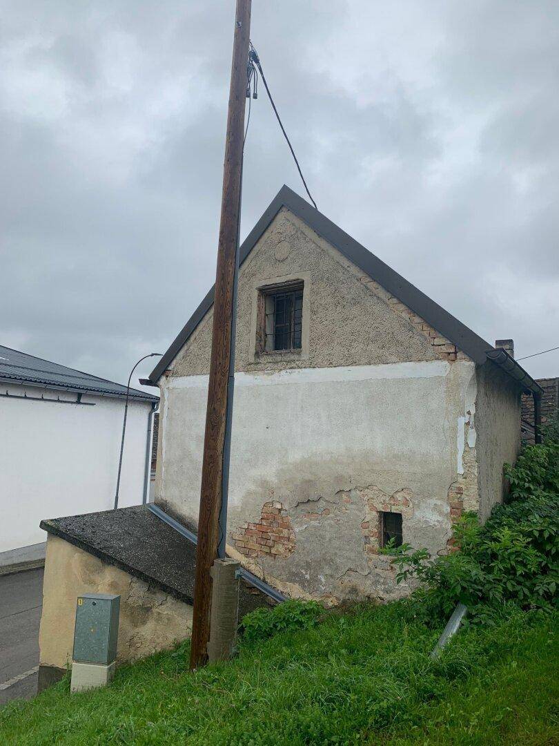 Seitenansicht eines älteren Hauses mit bröckelndem Putz und einem Giebelfenster, umgeben von Grün und einem Strommast.