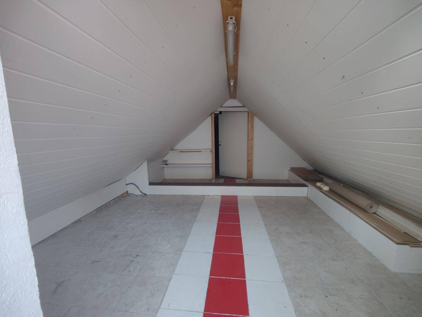 Ausgebauter Dachbodenraum mit schrägen Wänden, Holzverkleidung und Fliesenboden mit rotem Streifen.