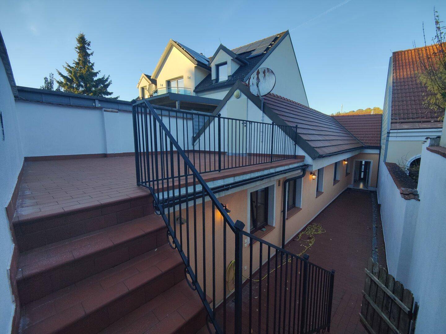 Große Außenterrasse mit roten Fliesen und Treppenzugang, umgeben von Gebäuden.