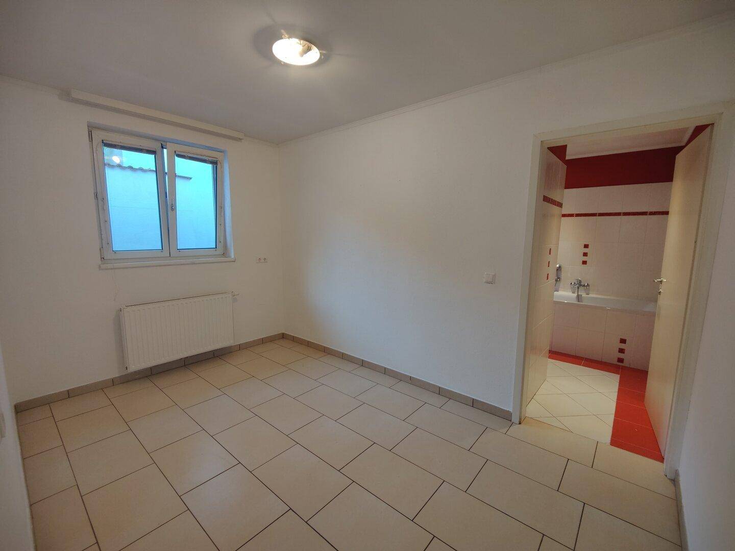 Schlafzimmer mit Fliesenboden und Fenster, angrenzend an ein Badezimmer mit roten Akzenten.
