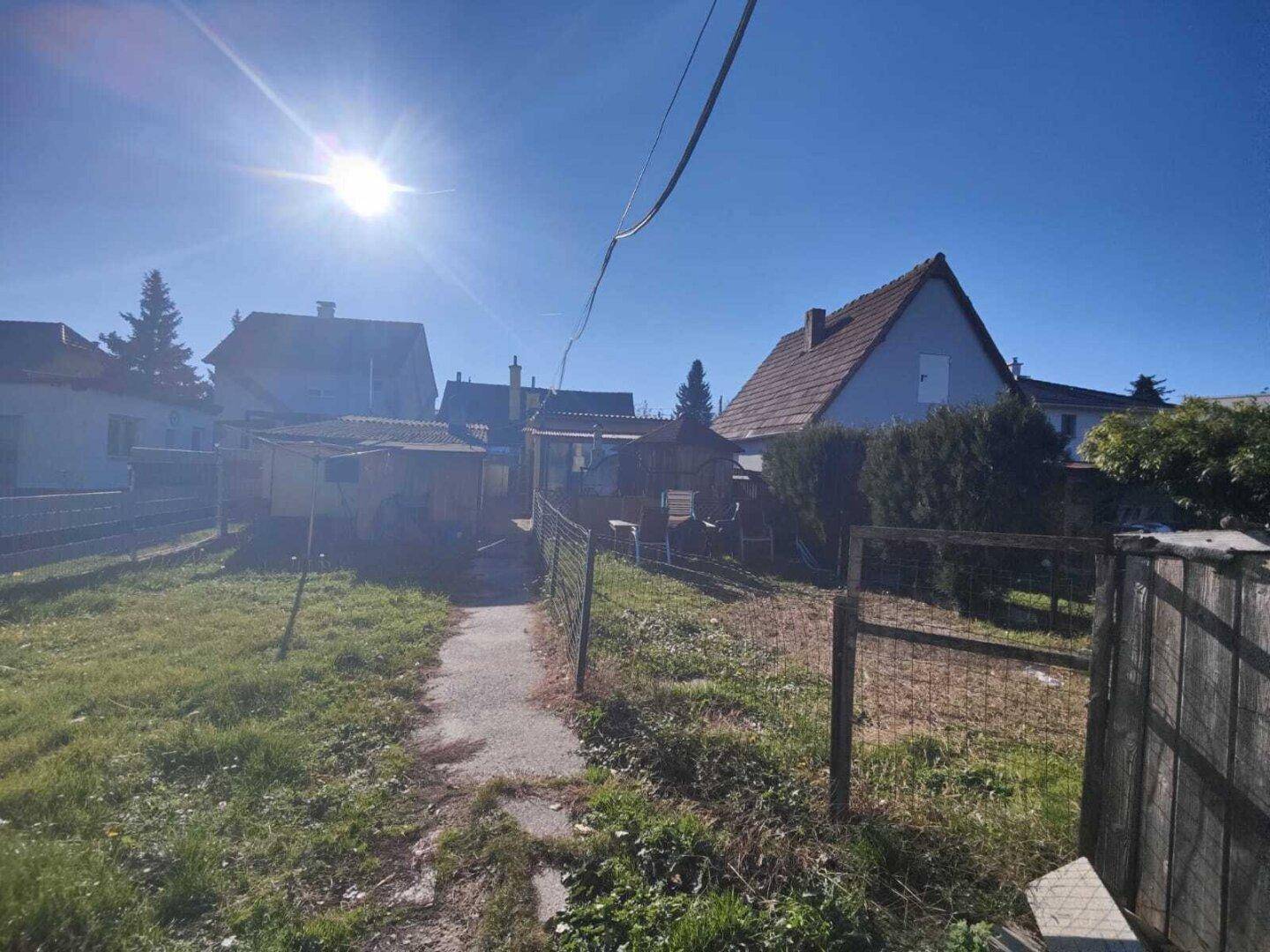 Gartenbereich mit Rasenfläche, einem Pfad und kleinen Nebengebäuden im Hintergrund unter strahlendem Sonnenschein.
