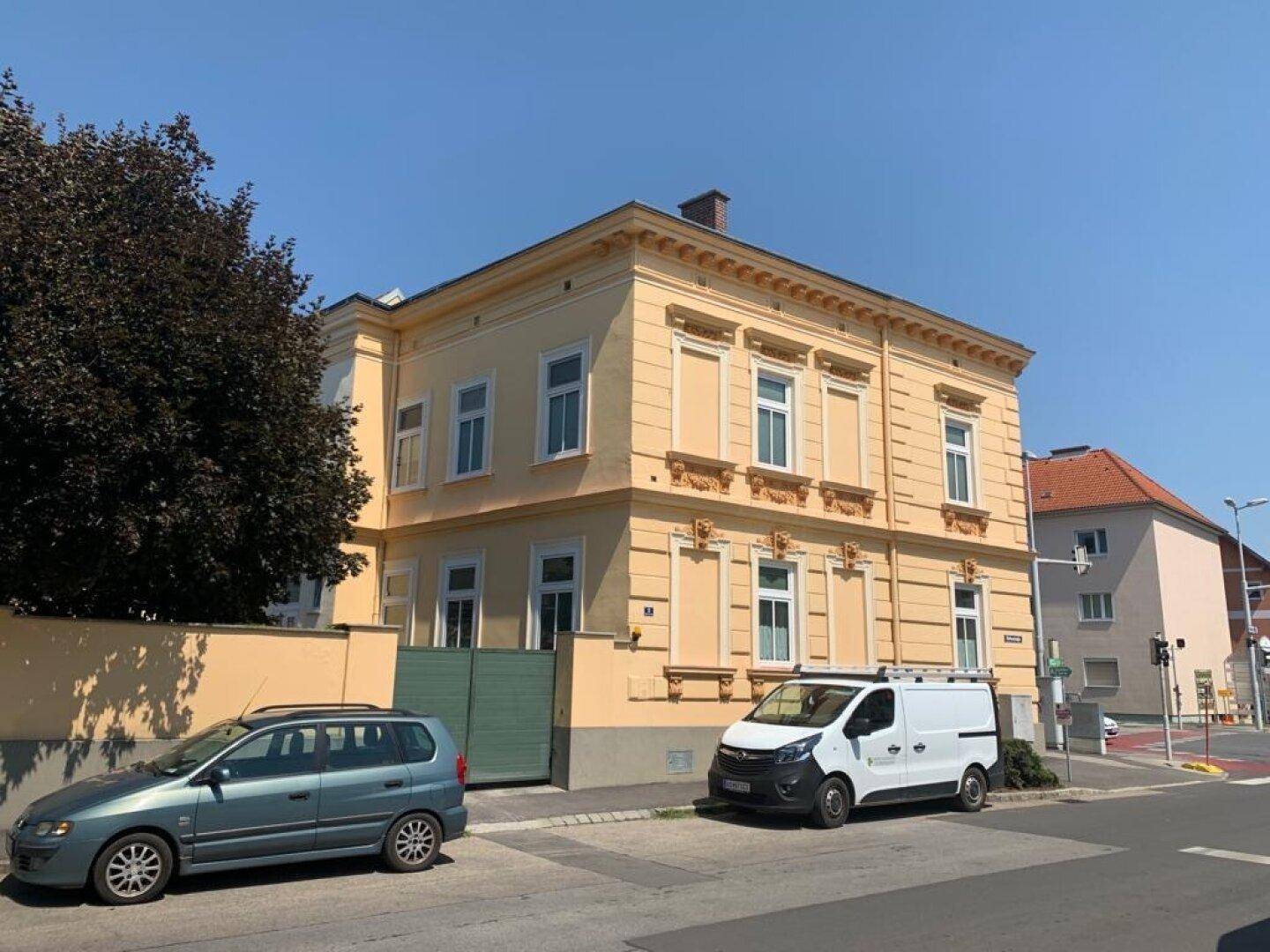 Seitenansicht des historischen Gebäudes mit gelber Fassade, grüner Toreinfahrt und geparkten Fahrzeugen.