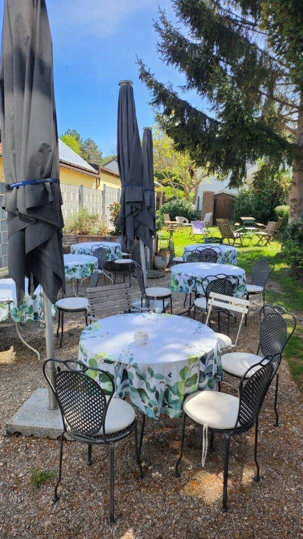 Gepflegter Gastgarten mit mehreren Tischen, Sonnenschirmen und Bäumen an einem sonnigen Tag.