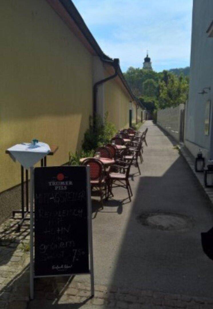 Schmaler Außenbereich mit Tischen und Stühlen entlang einer Gasse, mit Blick auf einen Kirchturm.