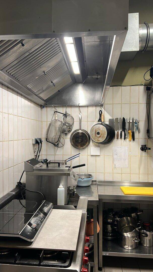 Gewerbliche Küche mit großer Dunstabzugshaube, Induktionskochfeld und Utensilien an der Wand.
