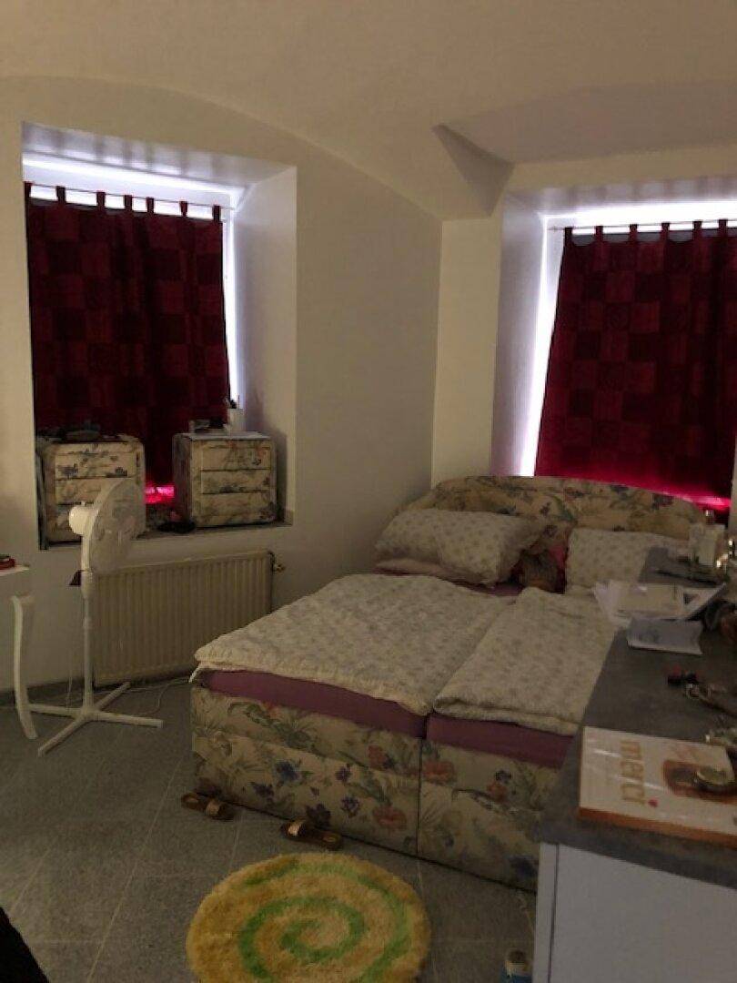 Schlafzimmer mit Bett, zwei Fenstern mit roten Vorhängen und grauem Fliesenboden.