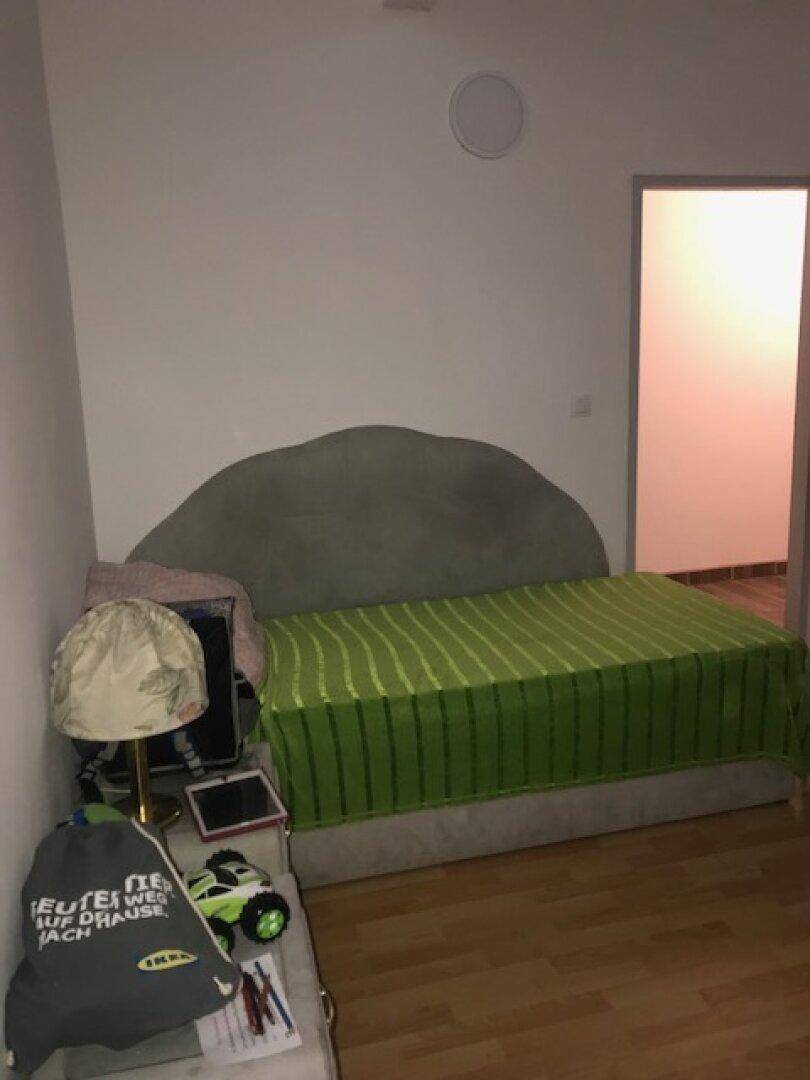 Schlafzimmer mit Bett, Nachttischlampe und hellem Laminatboden, mit grün gestreifter Bettdecke.