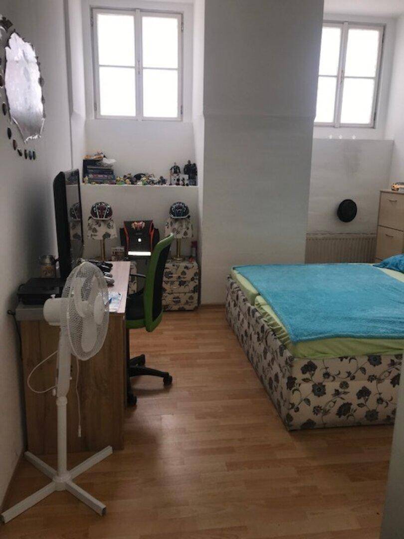 Schlafzimmer mit Bett, zwei Fenstern, Schreibtisch, Fernseher und hellem Laminatboden.