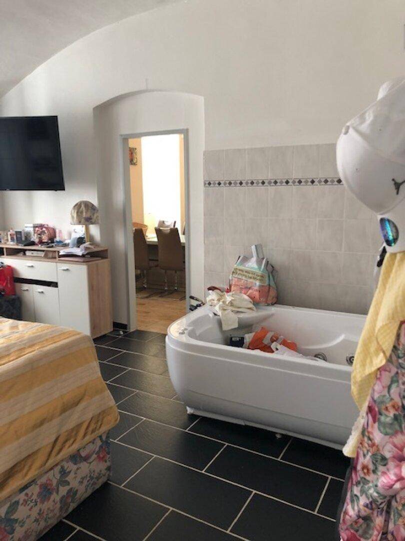 Schlafzimmer mit Bett, Fernseher und einer Badewanne im Raum, auf schwarzem Fliesenboden.