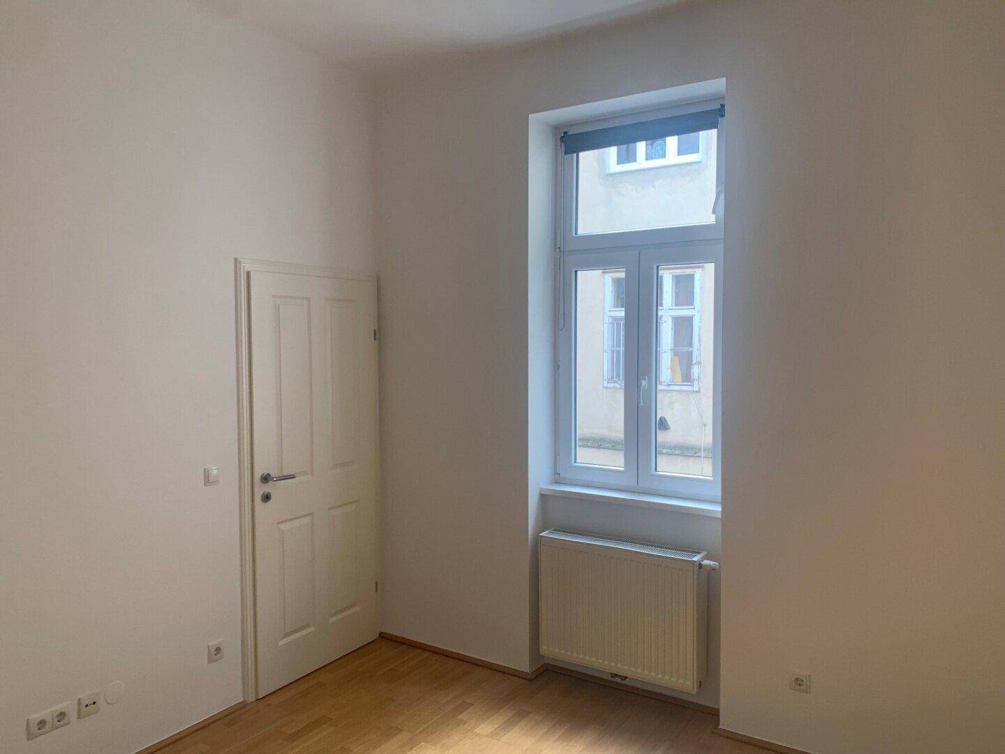 Leeres Schlafzimmer mit Holzboden, Fenster und weißer Tür.