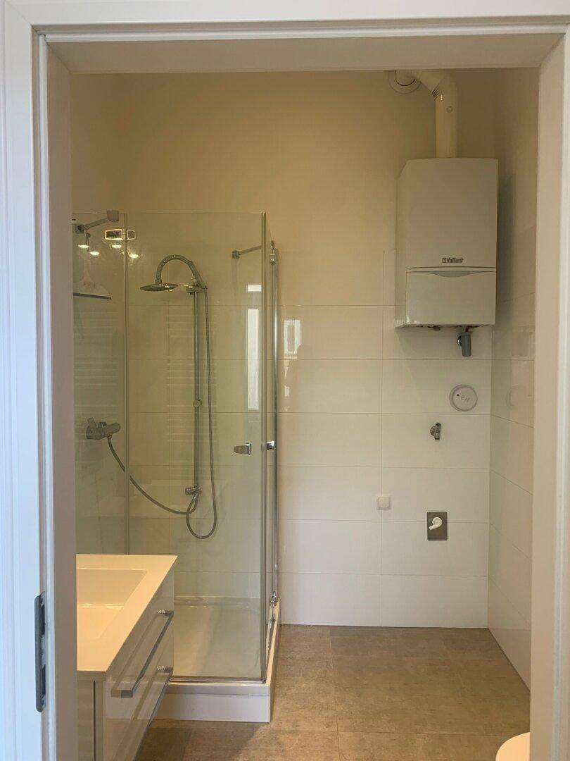 Helles Badezimmer mit Duschkabine, Waschbecken und wandmontiertem Warmwasserbereiter.