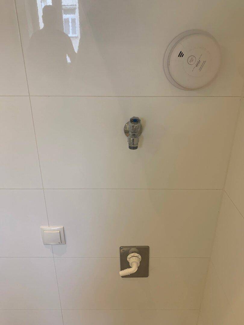 Detailansicht der weißen Fliesenwand mit Rauchmelder und Wasseranschlüssen.