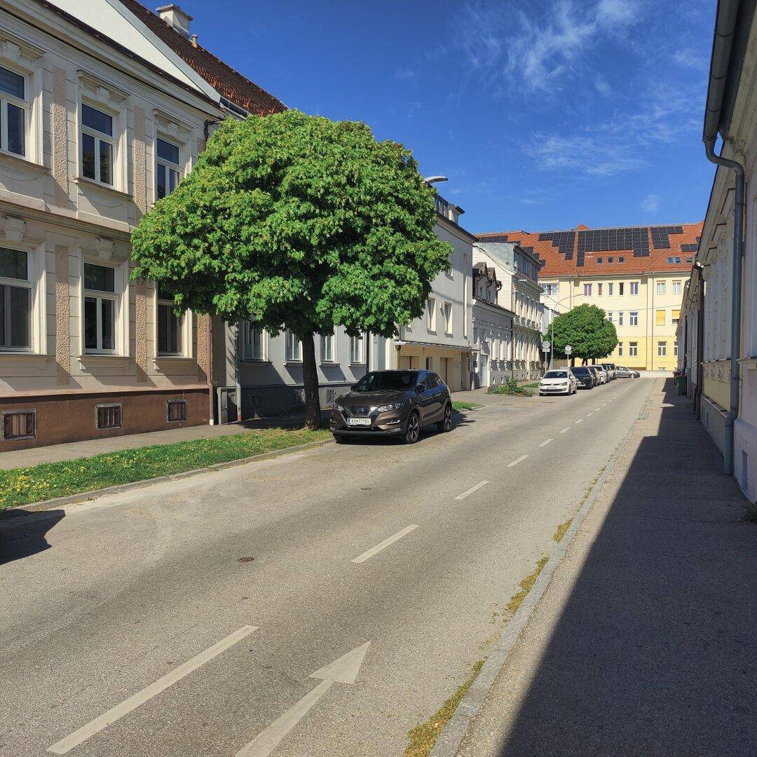 Sonnige Wohnstraße mit parkenden Autos, Bäumen und Gebäuden mit roten Dächern im Hintergrund.