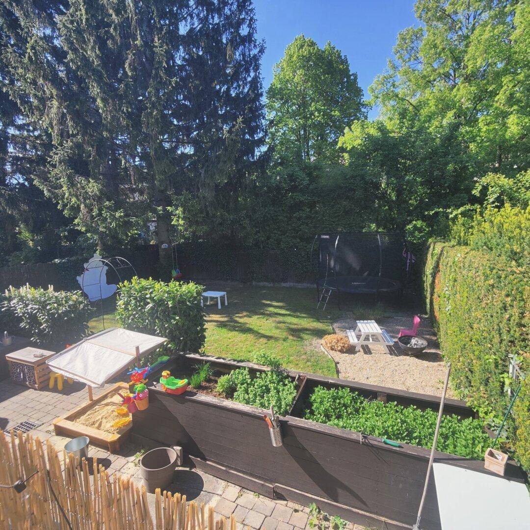 Gepflegter Garten mit Hochbeeten, Rasenfläche, Trampolin und hohen Bäumen im Hintergrund.