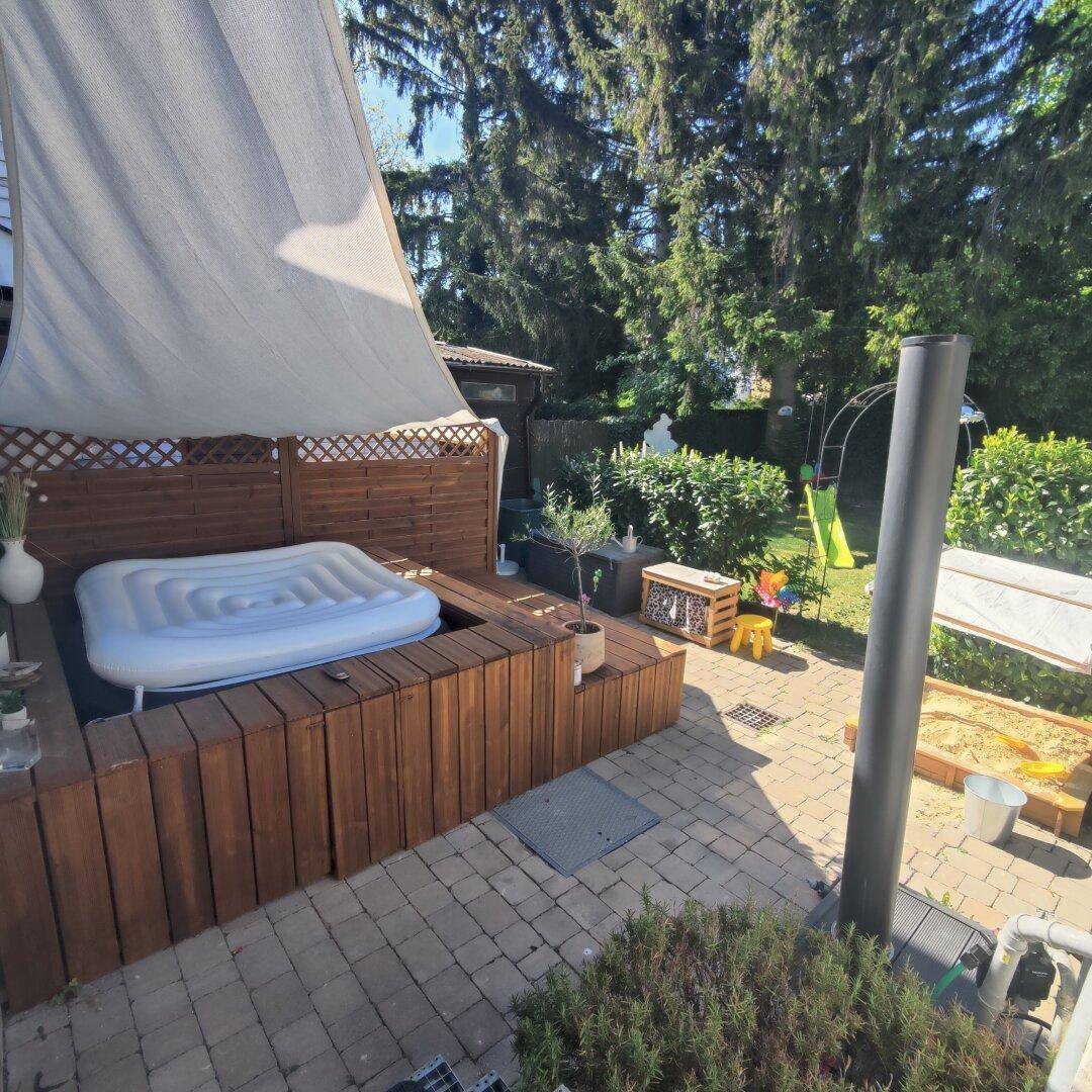 Gemütlicher Terrassenbereich mit Whirlpool, Holzverkleidung und Sonnensegel, umgeben von Grün.