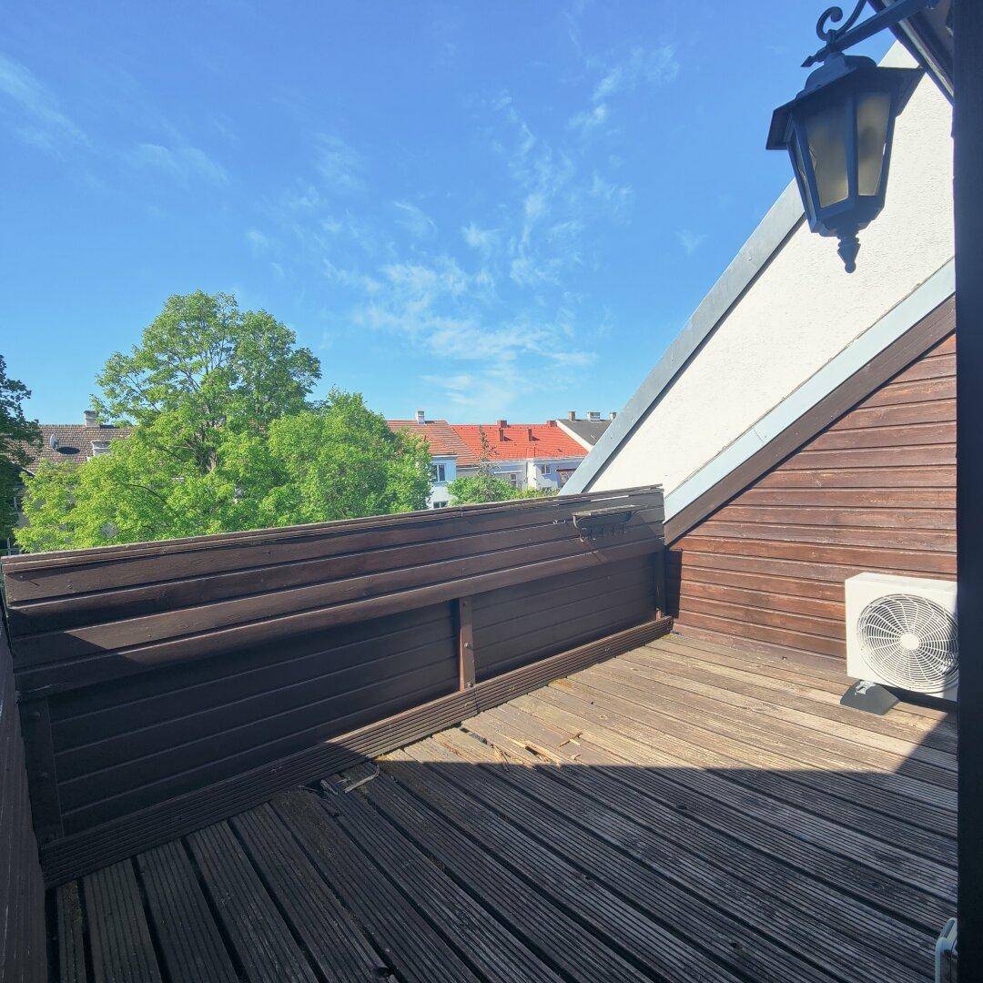 Holzbalkon mit Klimaanlage und Blick über Dächer und grüne Bäume an einem sonnigen Tag.