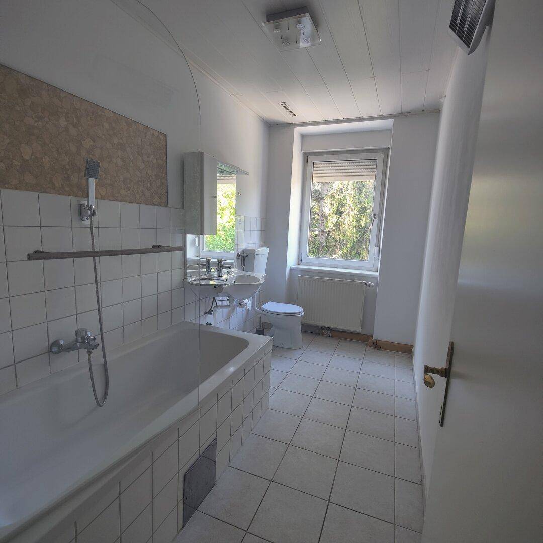 Helles Badezimmer mit Badewanne, WC, Waschbecken und Fenster, ausgestattet mit weißen Fliesen.