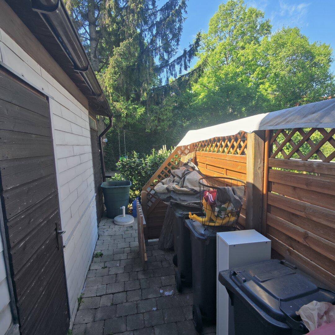Seitlicher Gartenbereich mit Mülltonnen, einem Schuppen und viel Grün, umgeben von Bäumen.