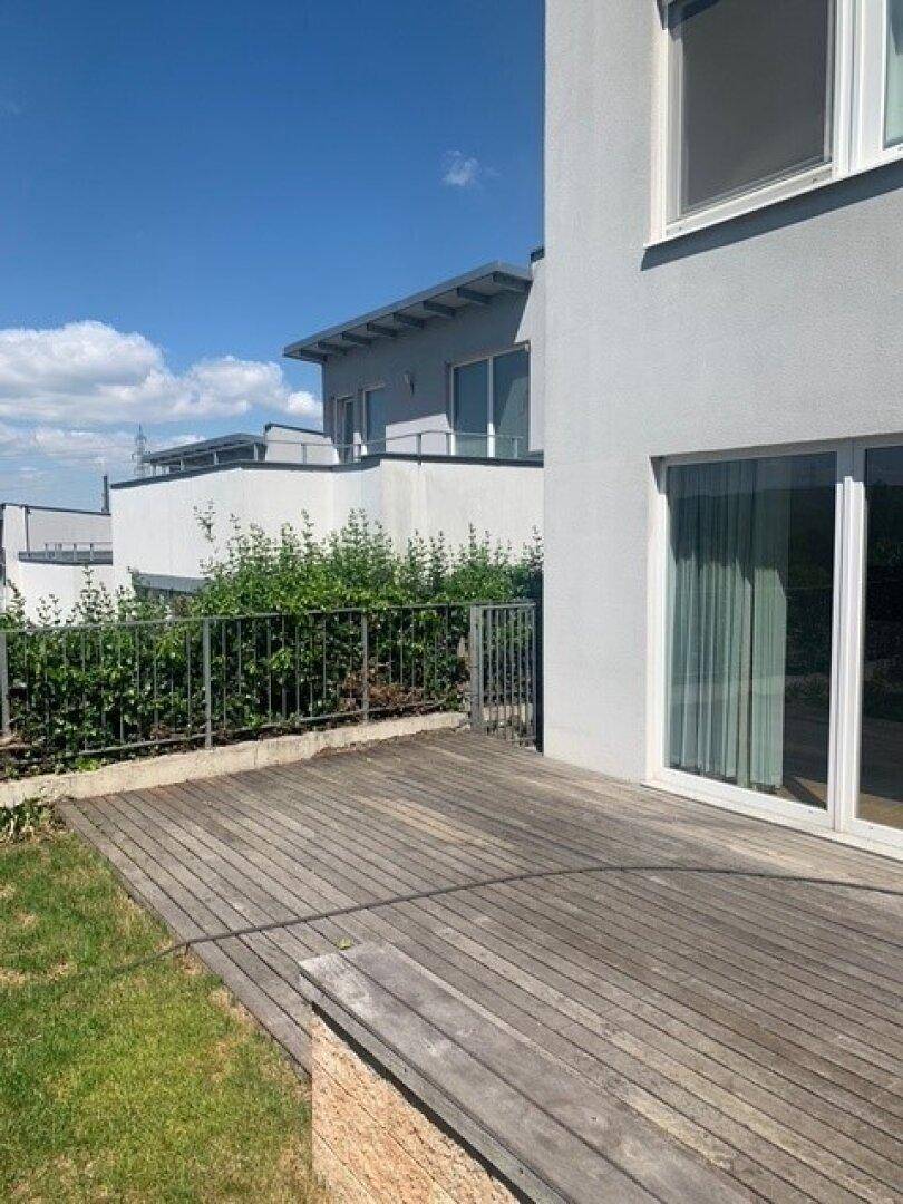 Seitliche Ansicht des Gebäudes mit Holzterrasse und einer grünen Hecke als Abgrenzung.