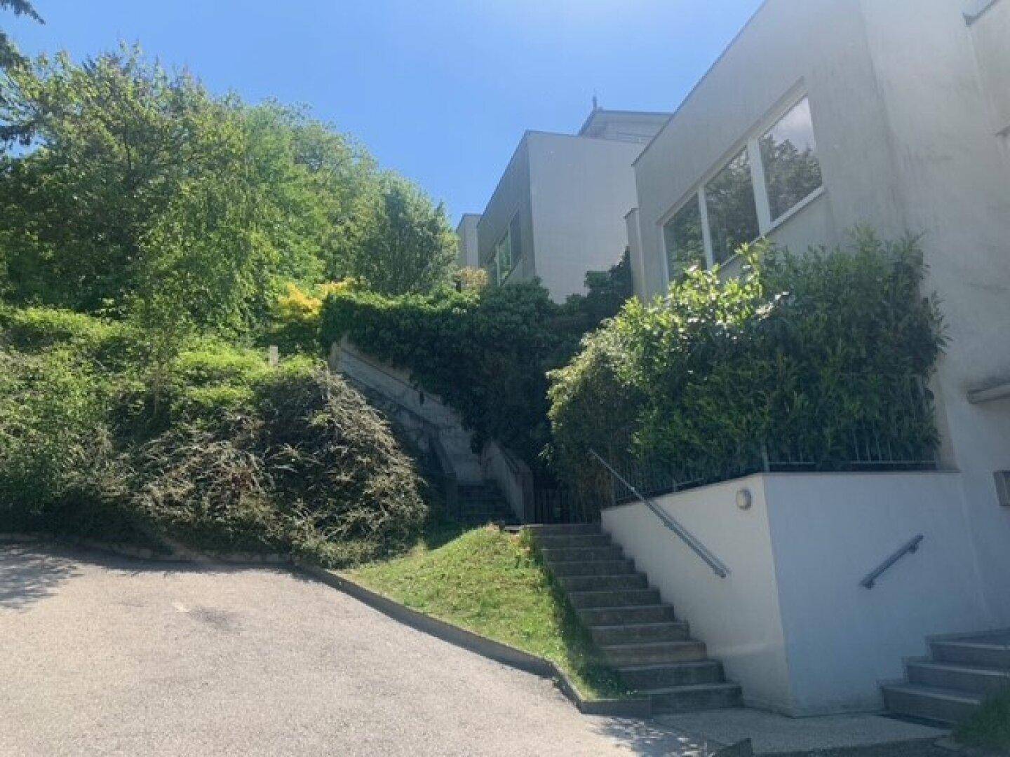 Moderne Gebäudefassade auf einem Hanggrundstück mit mehreren Ebenen und üppiger Bepflanzung.