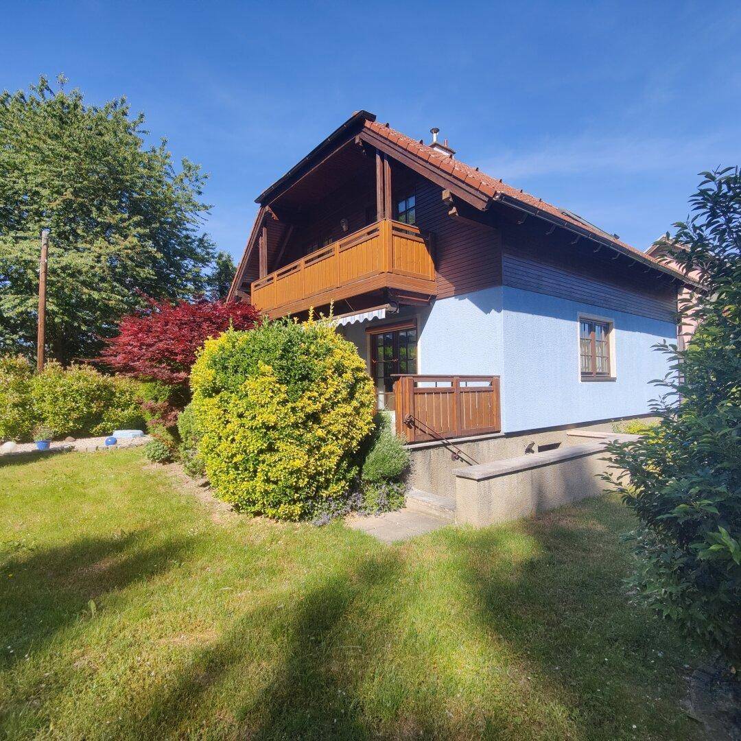 Seitenansicht des Hauses mit blauer Fassade, Holzelementen und einem Balkon, umgeben von Grünflächen.