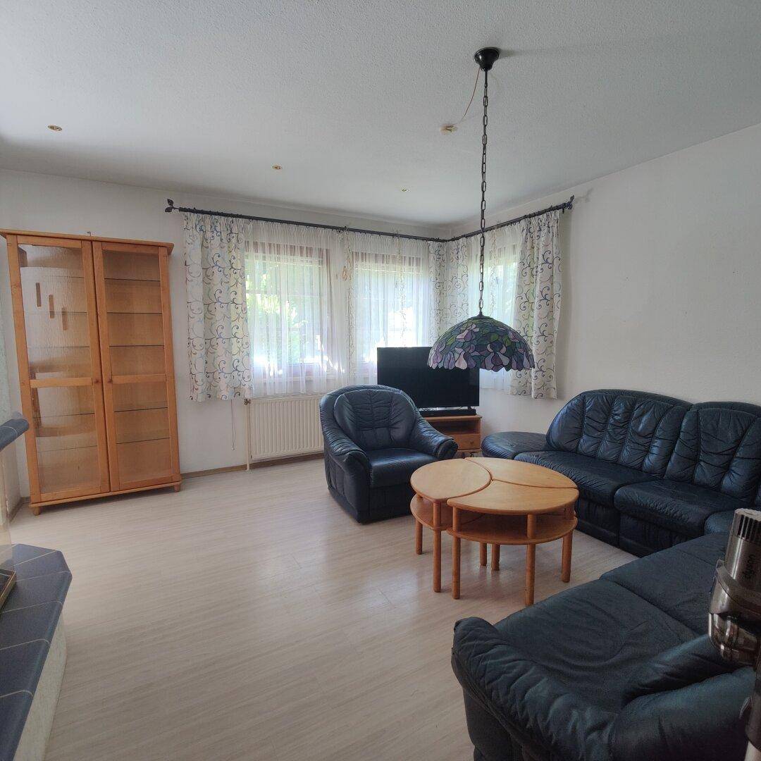 Gemütliches Wohnzimmer mit dunklen Ledersofas, Holztisch und großen Fenstern.