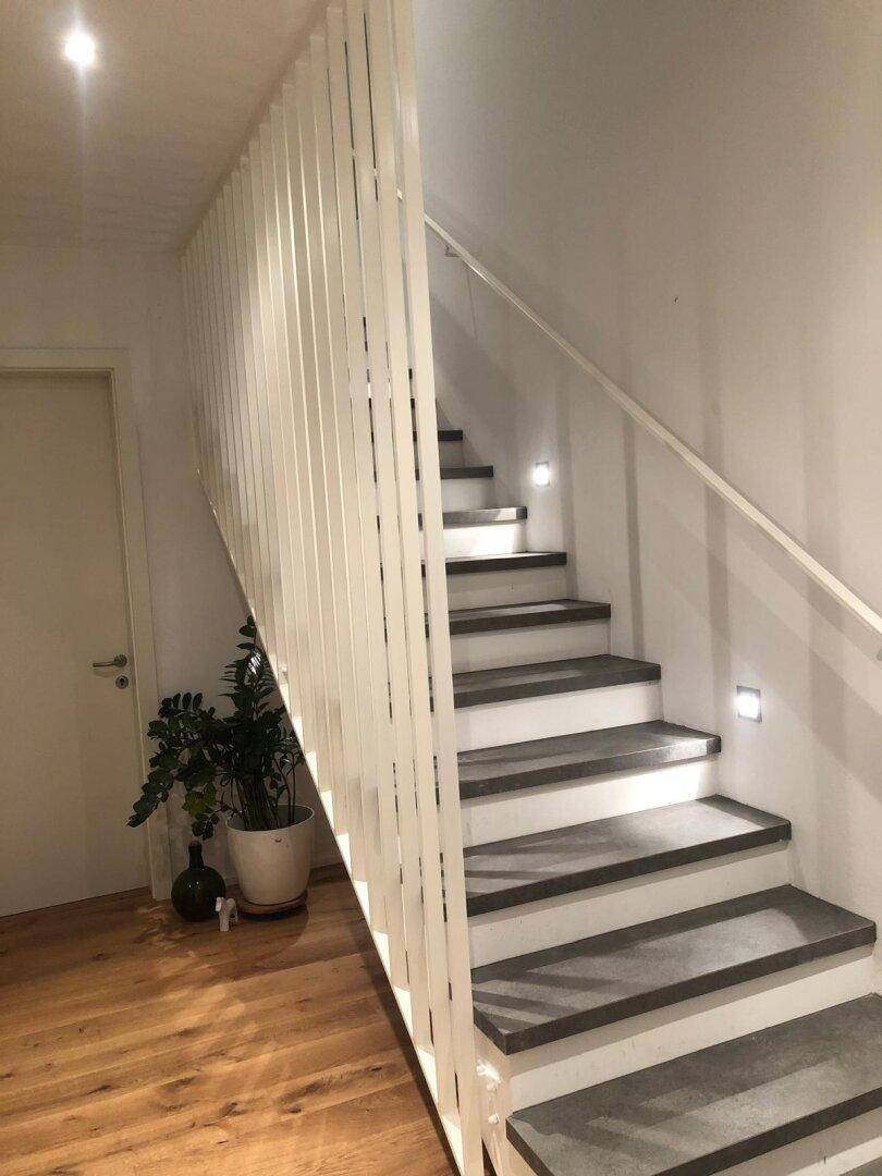 Moderner Treppenaufgang mit dunklen Stufen, weißem Geländer und indirekter Beleuchtung.