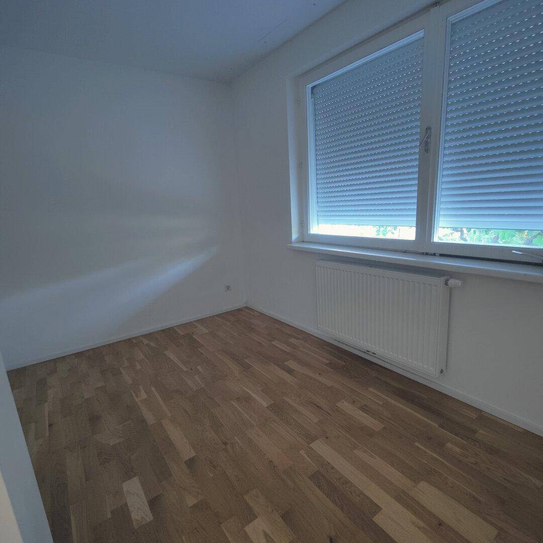 Helles Schlafzimmer mit Holzparkettboden, Fenster mit Rollladen und Heizkörper unter dem Fenster.