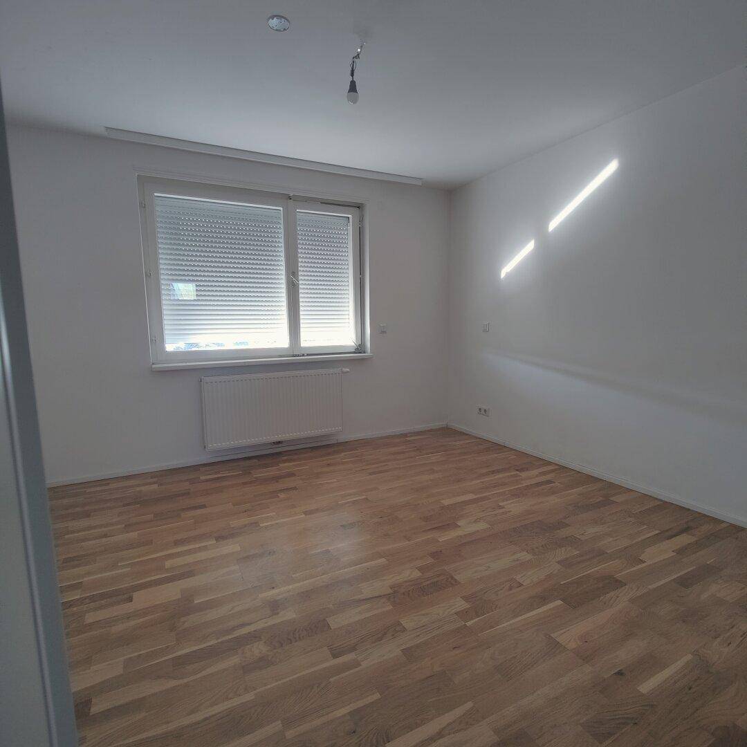 Geräumiges Schlafzimmer mit Holzparkettboden, großem Fenster mit Rollladen und viel Tageslicht.