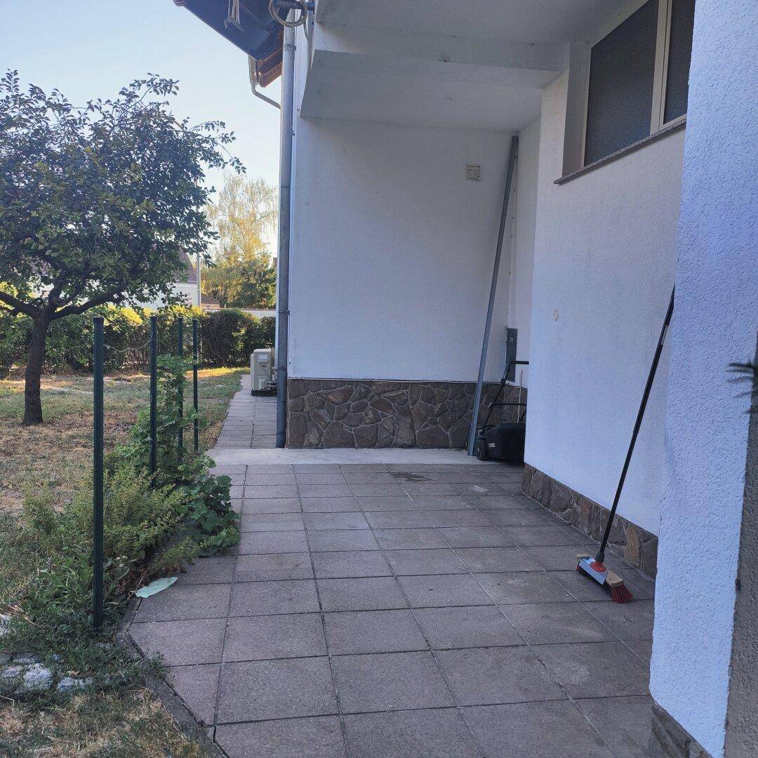 Gepflasterter Weg entlang des Hauses mit angrenzendem Gartenbereich und Bäumen.