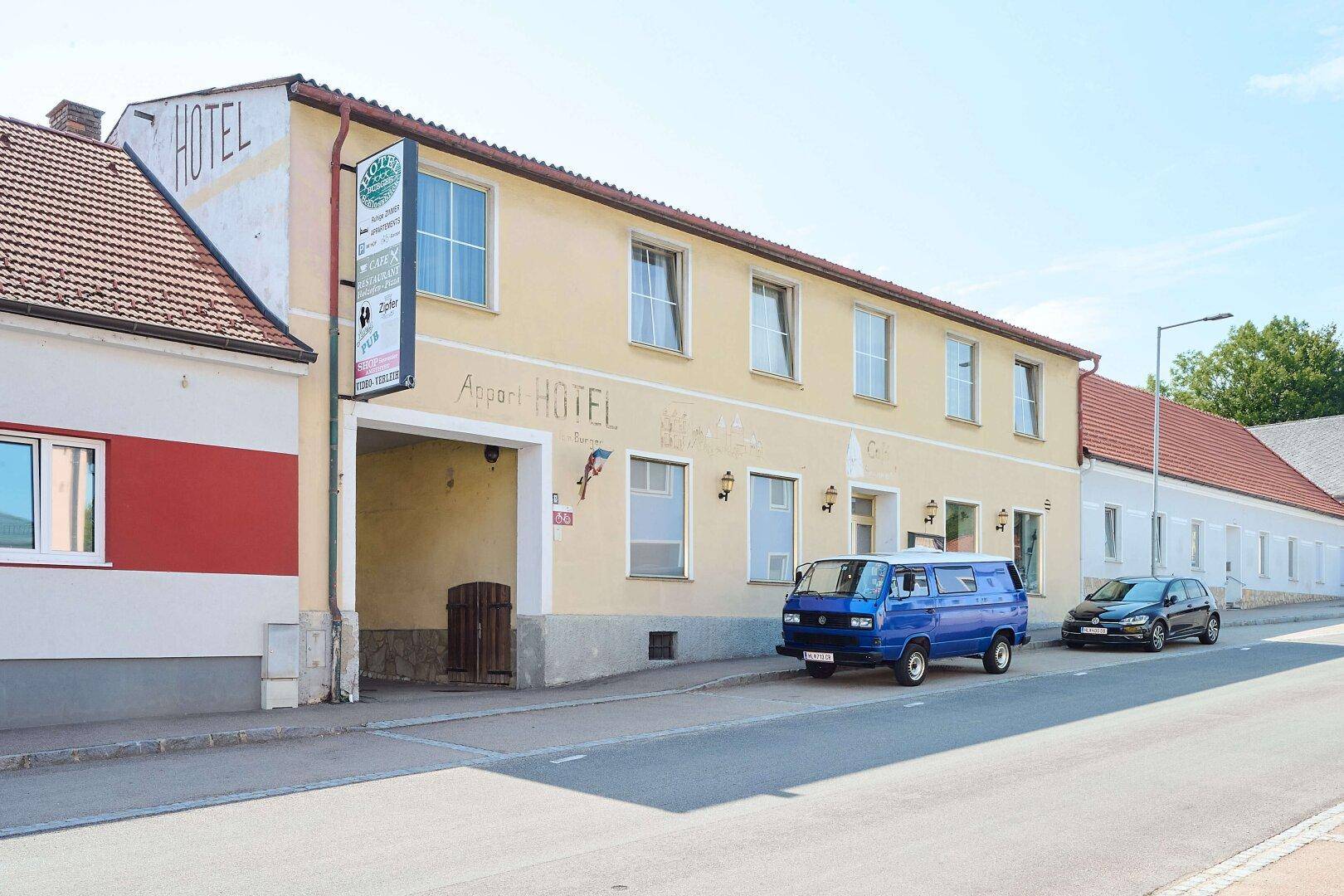 Ansicht des Hotels von der Straße mit geparkten Fahrzeugen und dem Schriftzug "HOTEL" an der Fassade.