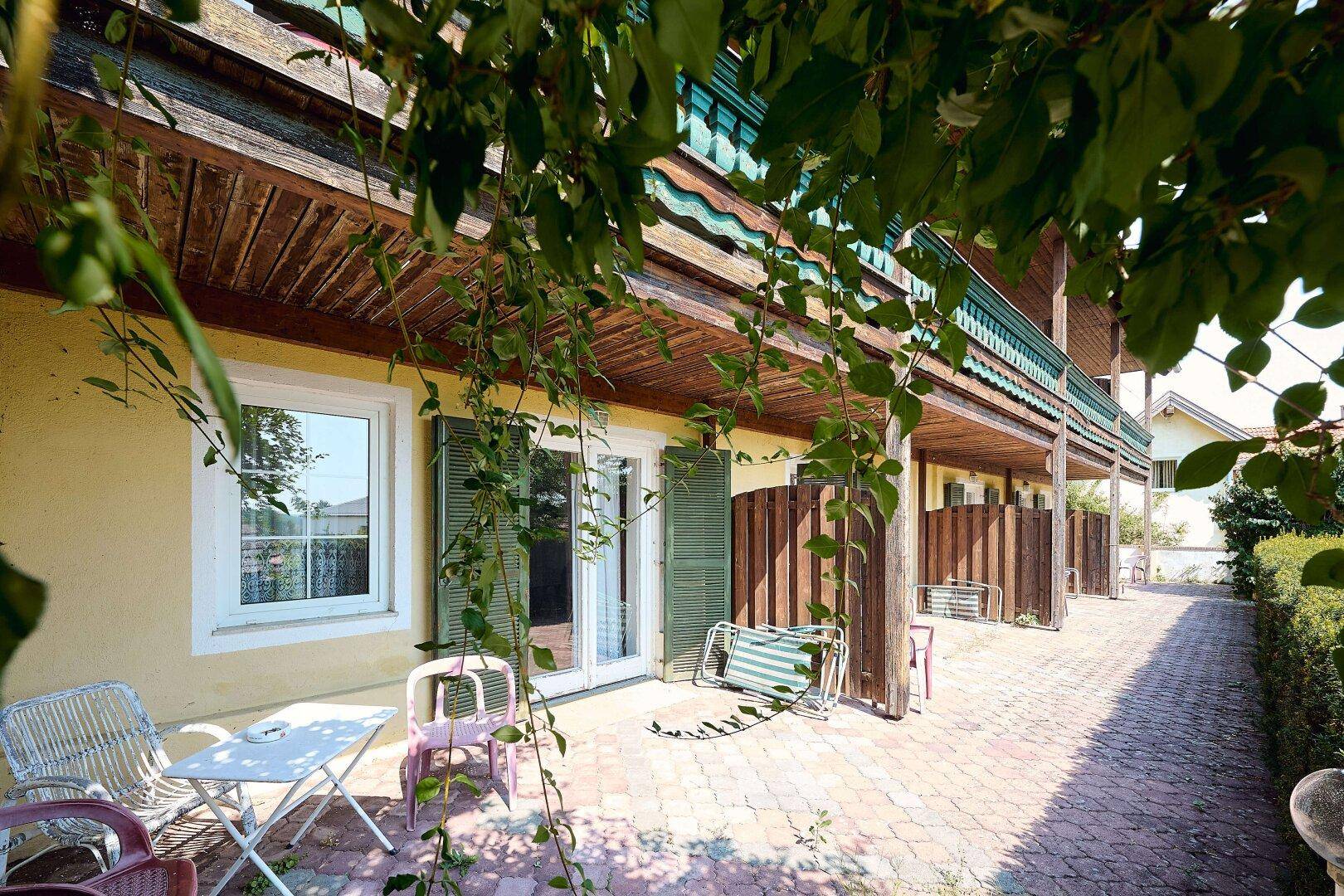 Eine gepflasterte Terrasse mit Gartenmöbeln und einem darüberliegenden Holzbalkon, umgeben von Grün.