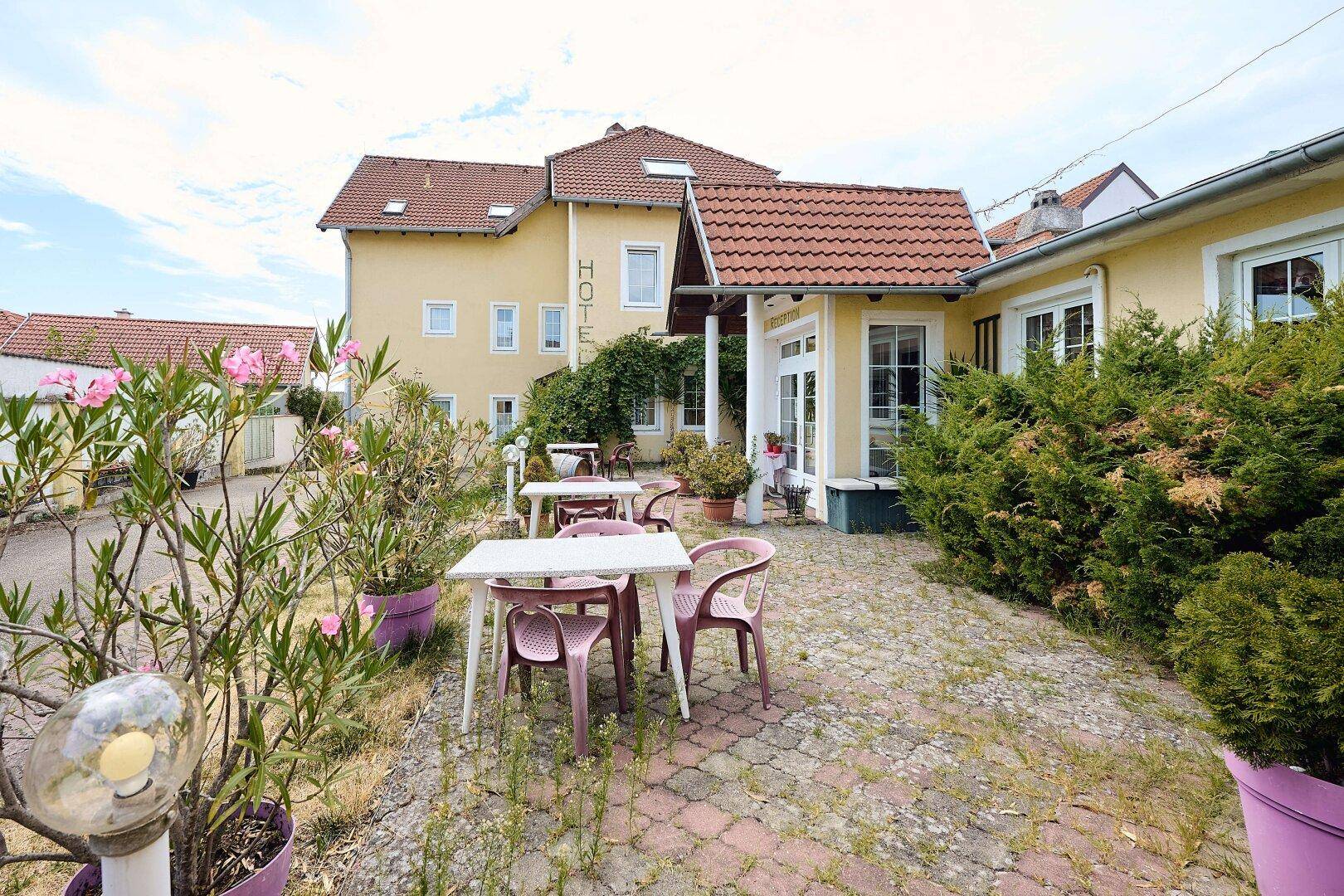 Ein Außenbereich mit Tischen und Stühlen, umgeben von blühenden Pflanzen und dem Hotelgebäude im Hintergrund.