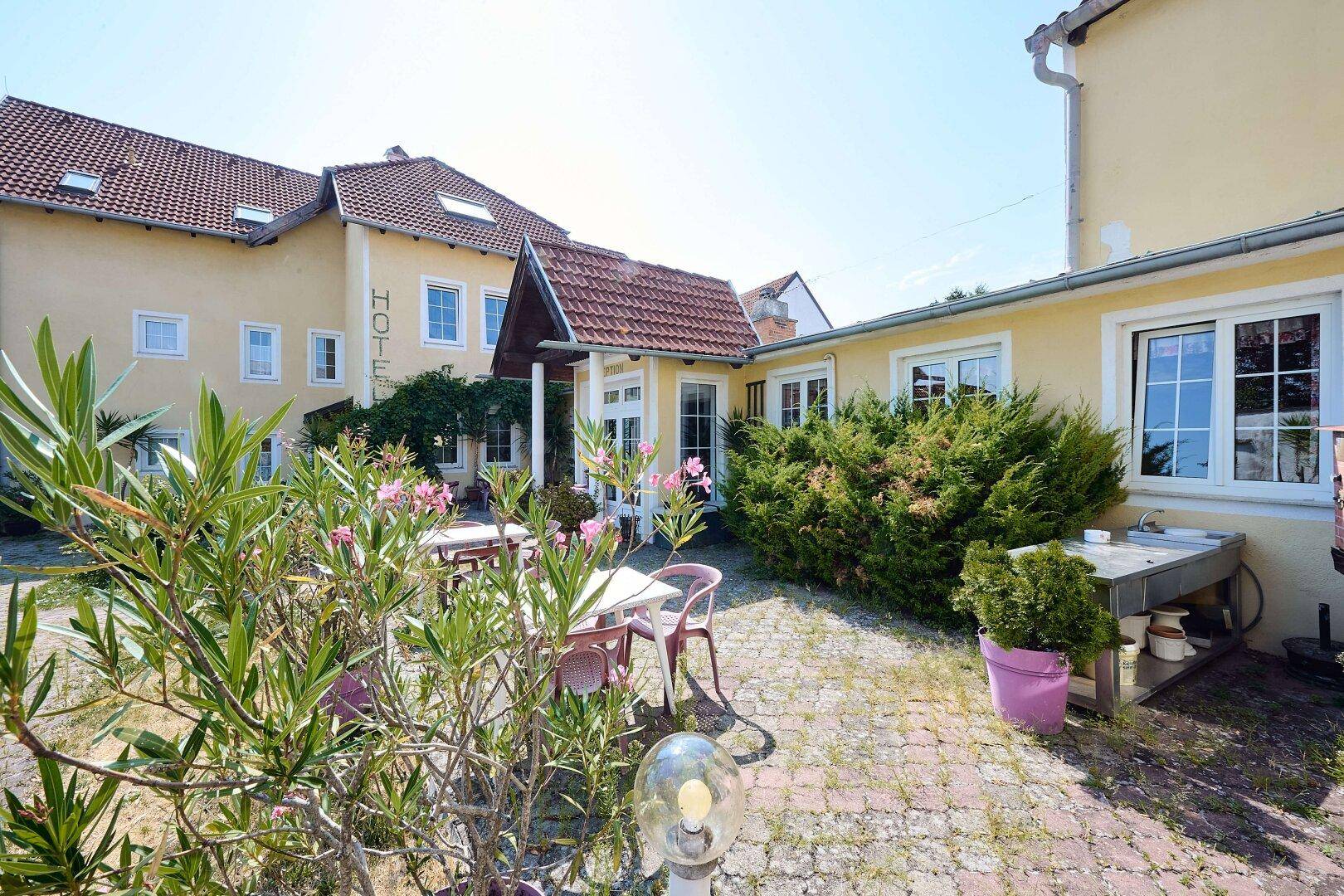 Ein Außenbereich mit Tischen und Stühlen, umgeben von blühenden Pflanzen und dem Hotelgebäude im Hintergrund.