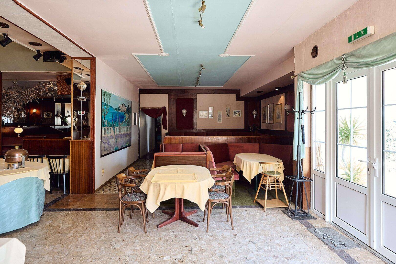 Eingangsbereich eines Gastraums mit Tischen, Stühlen und großen Fenstertüren zum Außenbereich.