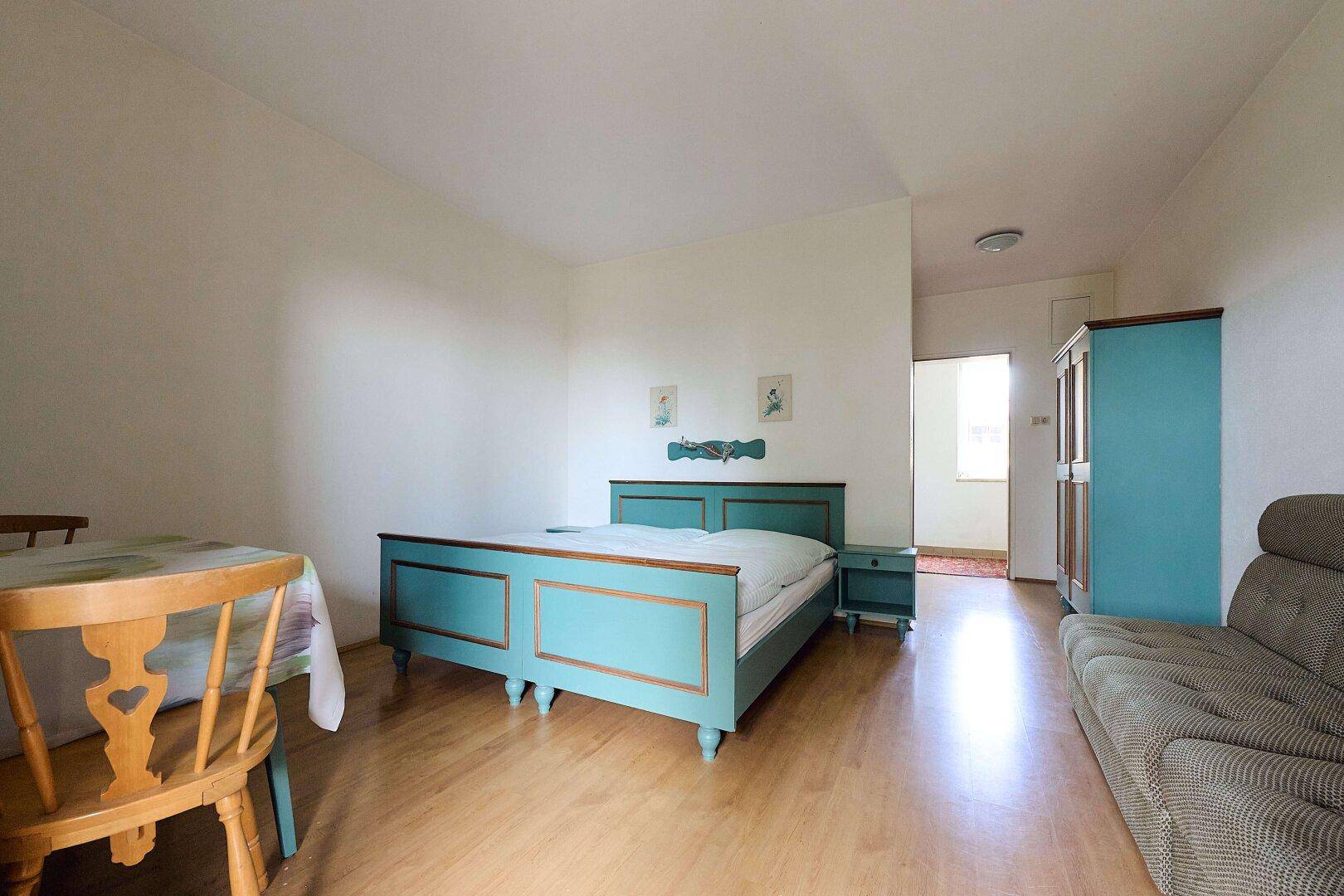 Schlafzimmer mit blau gestrichenem Bettrahmen, Sofa und einem Esstisch mit Stühlen.