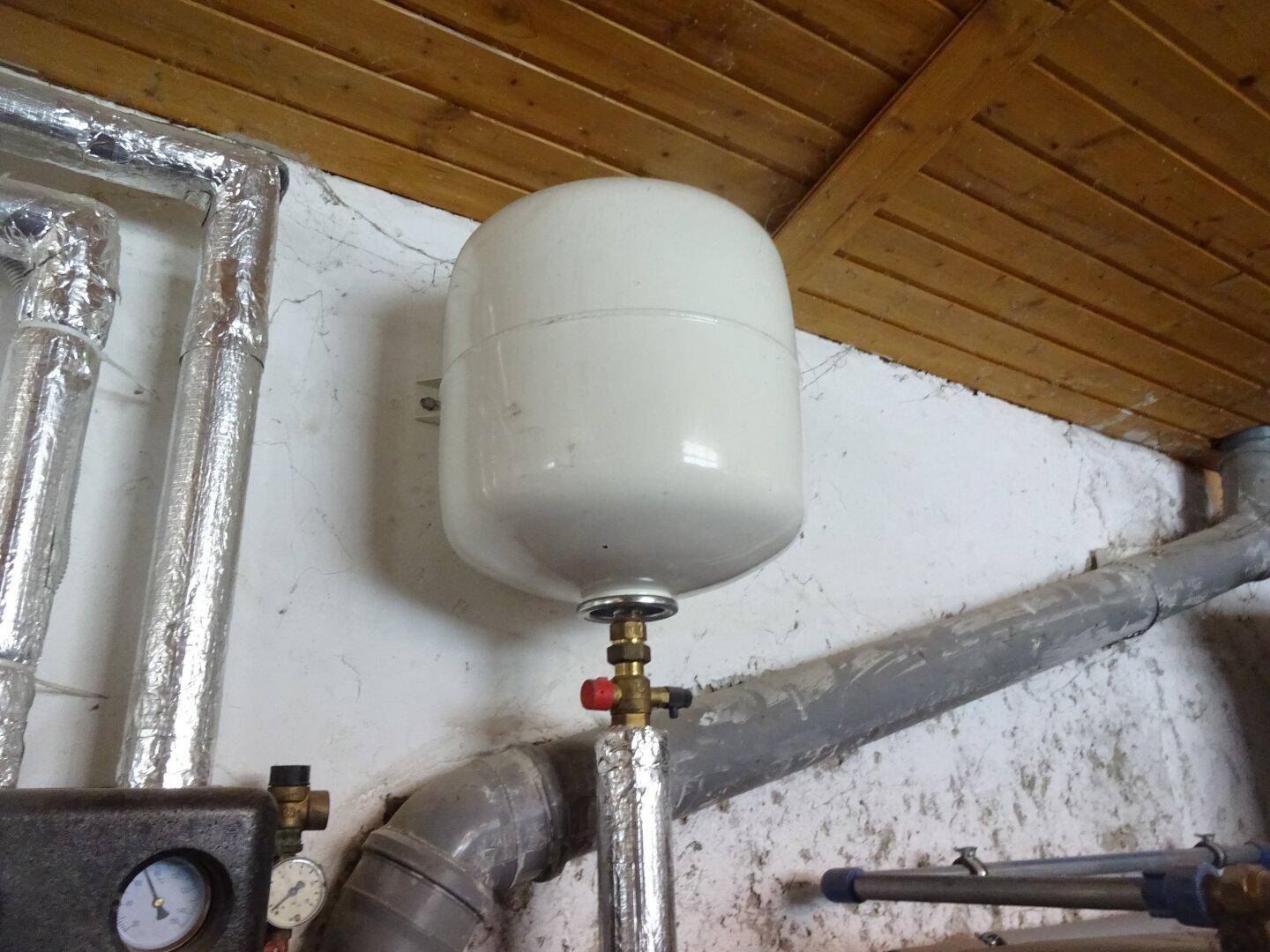 Weißes Ausdehnungsgefäß an der Wand in einem Heizungsraum mit Holzdecke und Rohren.