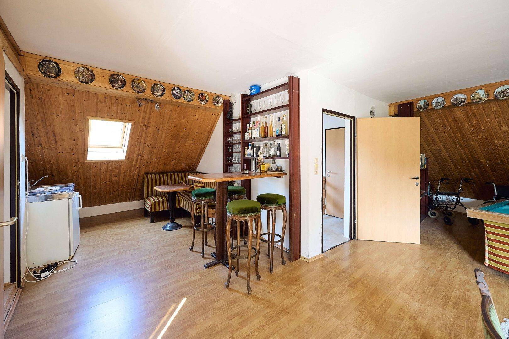 Dachbodenraum mit kleiner Bar, Sitzgelegenheiten und Holzverkleidung unter einem Dachfenster.