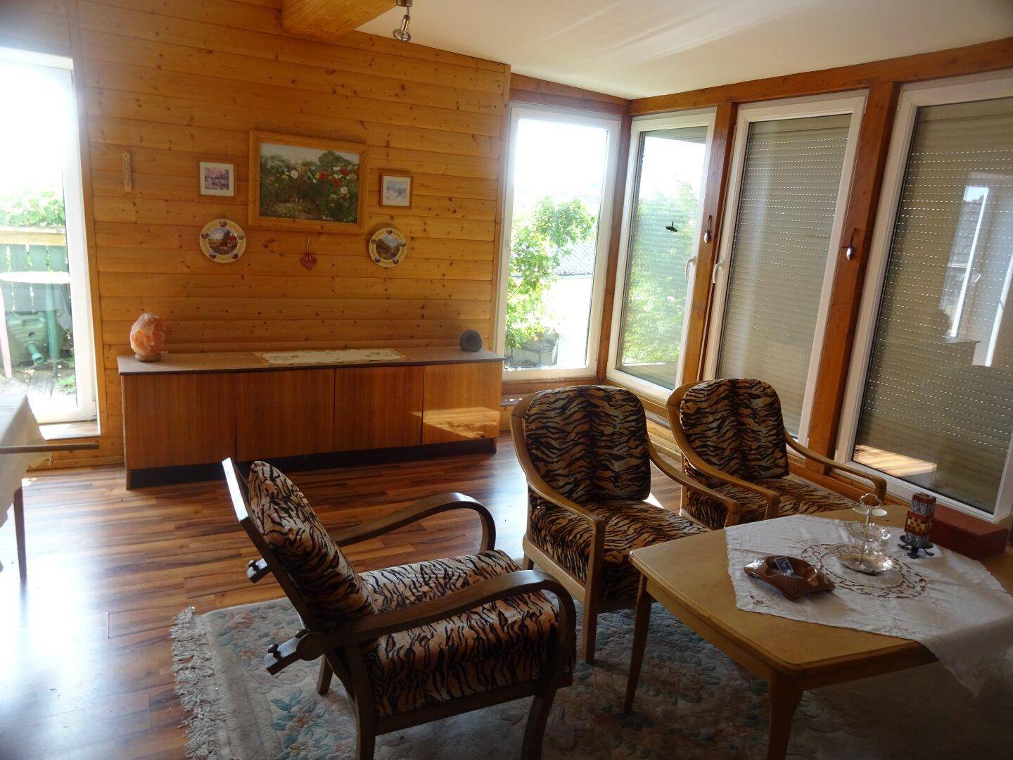 Helles Wohnzimmer mit Holzvertäfelung, großen Fenstern und gemusterten Sesseln.