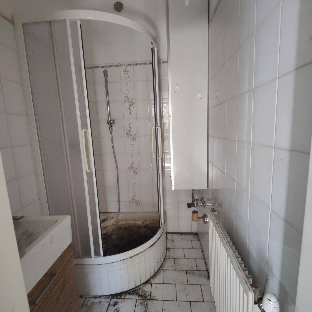 Kleines, altes Badezimmer mit Eckdusche, Waschbecken und Heizkörper, renovierungsbedürftig.