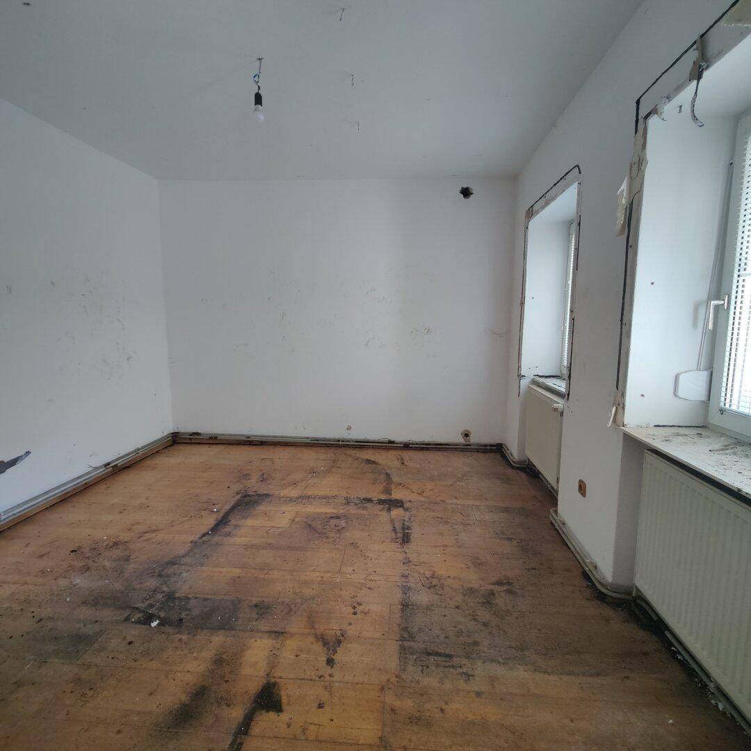 Leerer Raum mit abgenutztem Holzboden und zwei Fenstern, bereit für eine Renovierung.