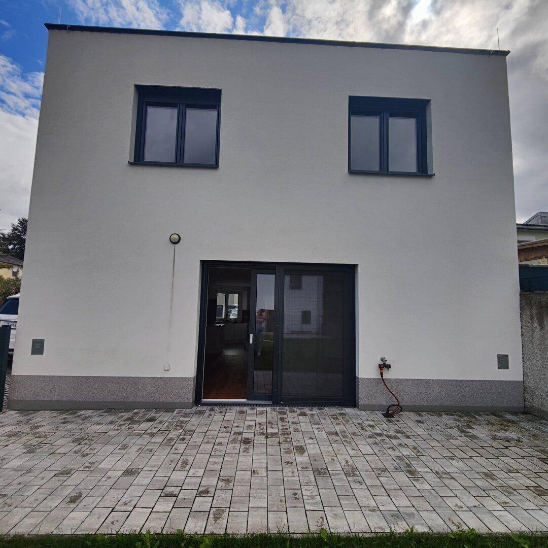 Rückseite des modernen Neubaus mit großer Glasschiebetür und gepflastertem Bereich.