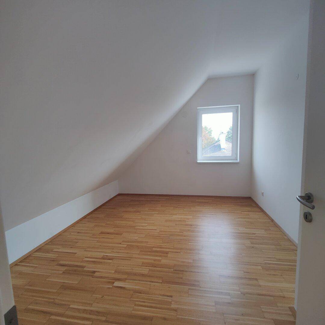 Helles Schlafzimmer mit Holzboden, Dachschräge und einem Fenster, das viel Tageslicht hereinlässt.