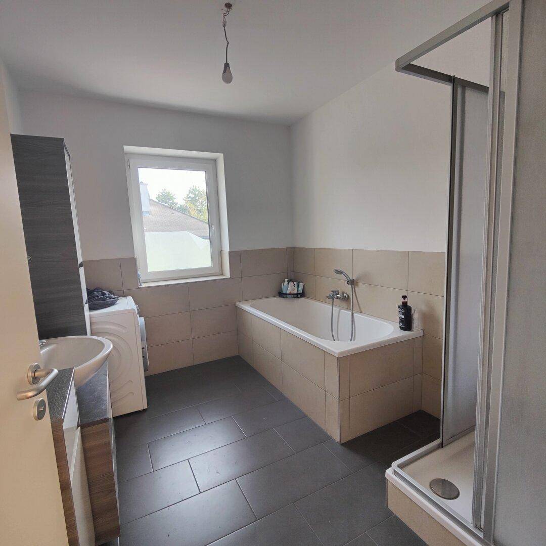 Badezimmer mit Badewanne, Duschkabine, Waschbecken und Fenster, ausgestattet mit Fliesenboden und Waschmaschine.
