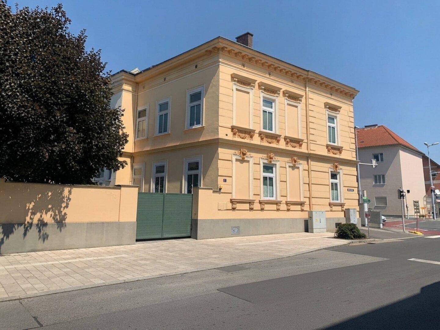 Seitenansicht des historischen gelben Gebäudes mit einer grünen Toreinfahrt und gepflastertem Bereich an der Straßenecke.