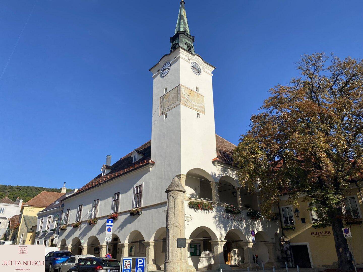 Historisches weißes Gebäude mit einem markanten Uhrturm und Arkaden, gelegen an einem sonnigen Stadtplatz.