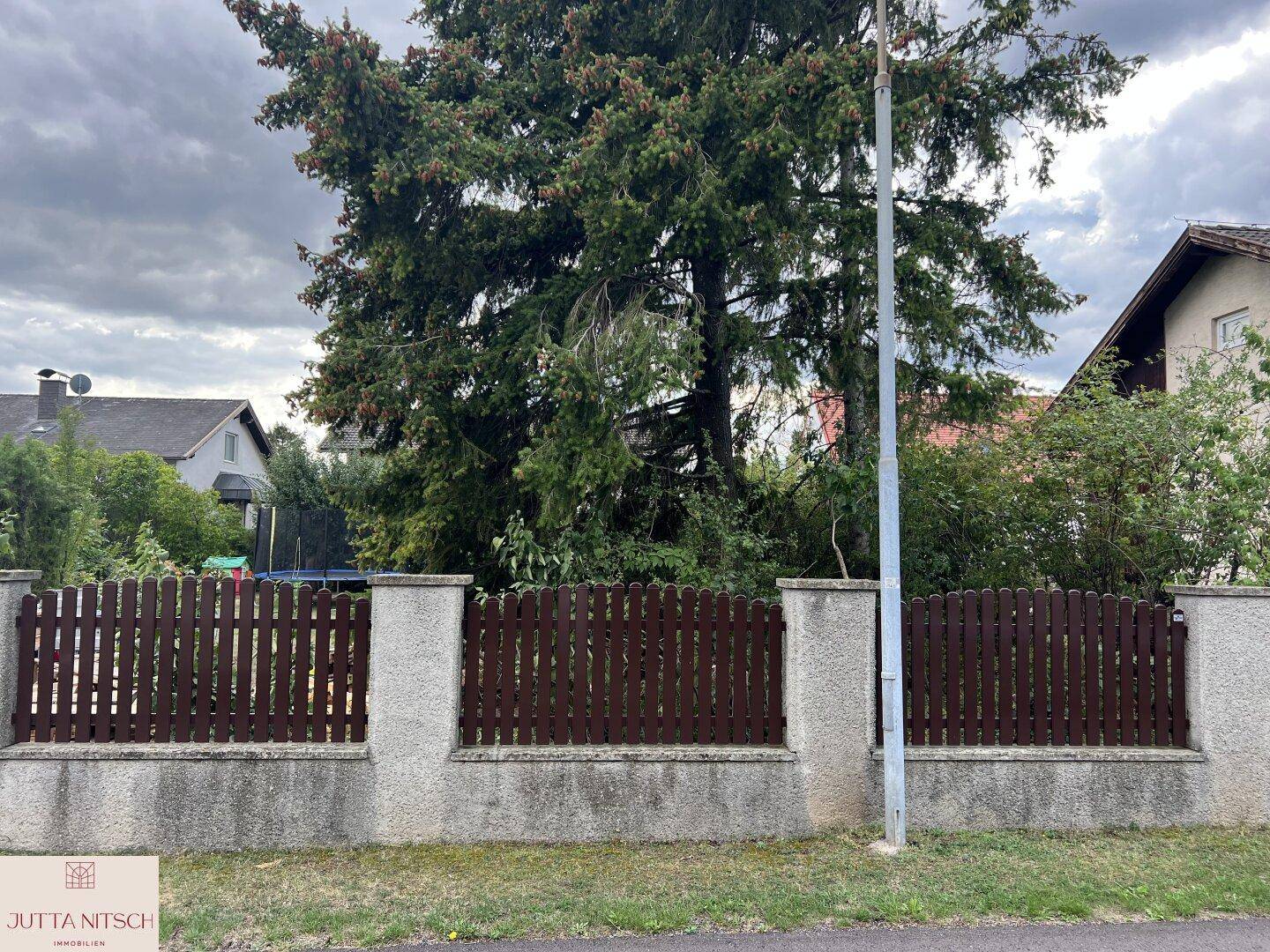 Robuster Holzzaun mit Betonpfosten entlang der Grundstücksgrenze, dahinter hohe Bäume.
