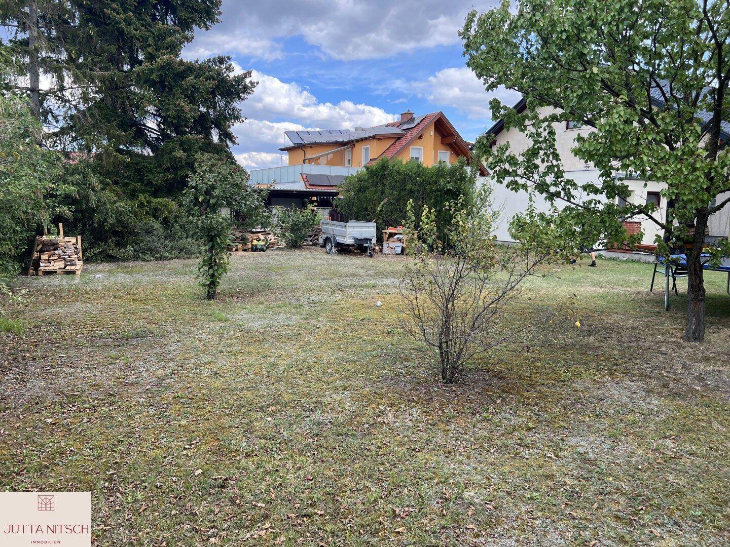 Gartenbereich mit Rasenfläche, Holzstapeln und einem Anhänger, umgeben von Vegetation.